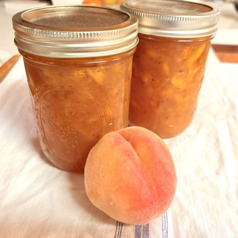 two pint jars of peach jam and a ripe peach sitting on a white kitchen towel