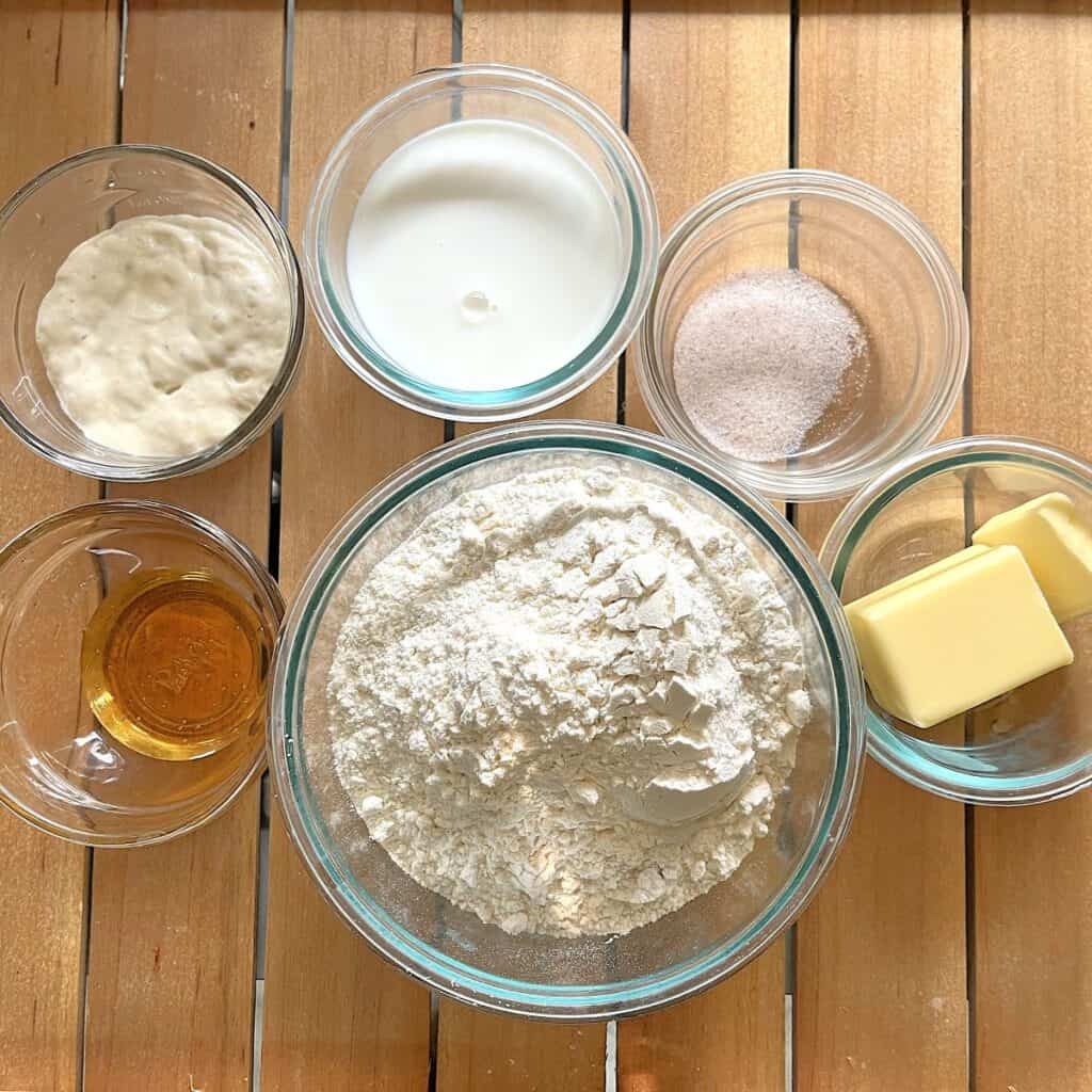 ingredients for sourdough rolls in glass dishes including sourdough starter, milk, salt, honey, flour, and butter