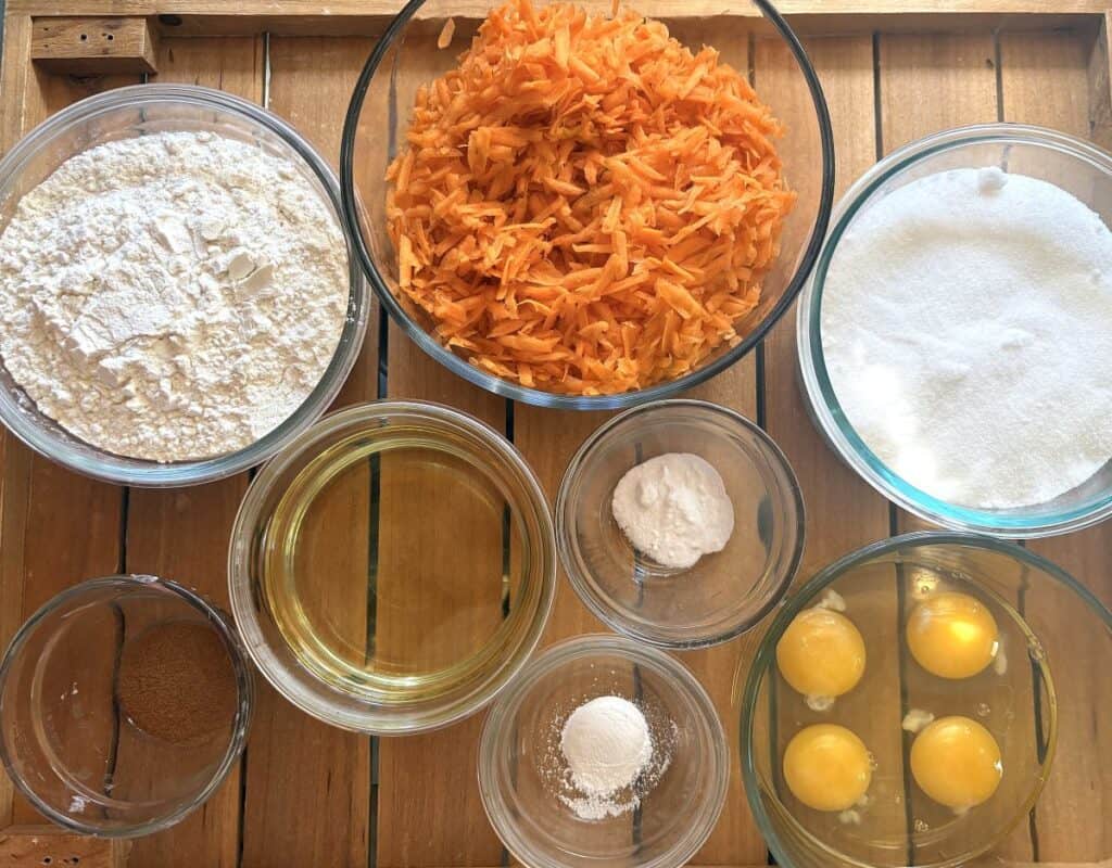 ingredients for carrot walnut cake including flour, shredded carrots, cinnamon, baking powder, baking soda, eggs, sugar, and cooking oil on a wooden tray